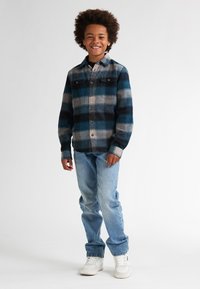 Camisa de cuadros de franela en azul, gris y negro con bolsillos con botones. Lucida con jeans azul claro y zapatillas blancas. Estilo casual.