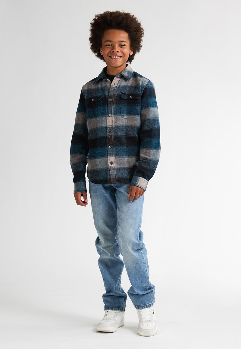 Camisa de cuadros de franela en azul, gris y negro con bolsillos con botones. Lucida con jeans azul claro y zapatillas blancas. Estilo casual.