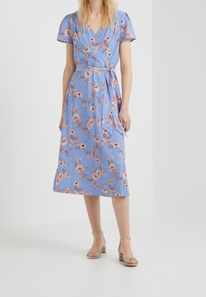 Kobieta w jasnoniebieskiej sukience w kwiaty na kopertę z krótkimi rękawami i beżowych sandałach na obcasie, stojąca na tle jednolitego białego tła.