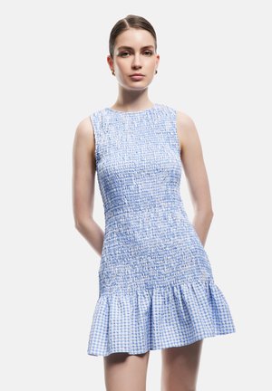 Giovane donna con capelli lisci pettinati all’indietro che indossa un mini vestito smockato senza maniche blu e bianco con orlo arricciato, in piedi davanti a uno sfondo semplice.