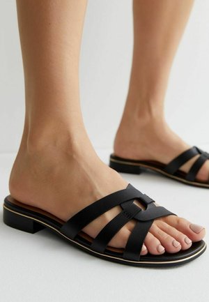 Füße, die schwarze flache Sandalen mit drei breiten Riemen tragen, von denen einer durch eine quadratische Schnalle gezogen ist, vor hellem Hintergrund.