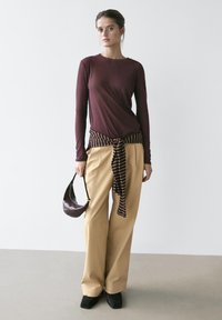 Femme portant un haut manches longues couleur bordeaux, un pantalon large beige, des chaussures noires, une chemise rayée nouée à la taille, tenant un petit sac à main sombre.