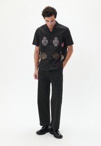 Camisa negra de manga corta con detallados bordados florales metálicos en dorado, plateado y rosa. Combinada con pantalones negros y zapatos brillantes.