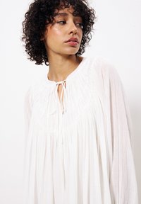 Weiße Bluse mit Rundhalsausschnitt, Kordelzug-Details und plissiertem Stoff. Lange, fließende Ärmel verleihen eine weiche Textur.