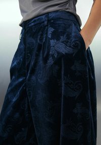 Close-up of dark blue velvet pants with paisley pattern, worn with a tucked gray shirt, hand placed in pocket at the hip.