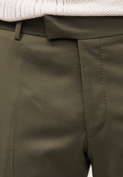 Κοντινό πλάνο στη μέση χακί παντελονιού με θηλιά για ζώνη και ένα λευκό υφασμάτινο πουλόβερ που έχει βγει μέσα.