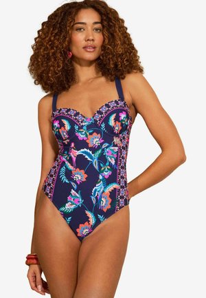 Vrouw met krullend haar die een marineblauw bloemend badpak met vogels en geometrische patronen draagt, poserend met één hand op haar heup.