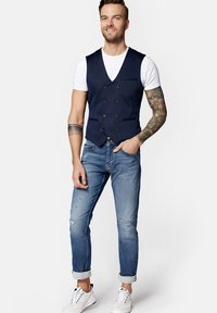 Granatowa kamizelka dwurzędowa na białym T-shircie, połączona z jasnoniebieskimi dżinsami z przetarciami i białymi sneakersami, z podwiniętymi mankietami.