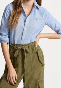 Ljusblå button-up skjorta med vikta ärmar, prydd med en liten broderad logotyp. Olivgröna högmidjade cargobyxor med knytband.