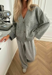 Szürke kötött kardigán mély V-nyakkal, gombos előlappal és zsebekkel, laza szürke sweatpants-szel párosítva. Texturált, laza otthoni viselet.