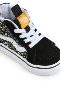 Svarta mockaskor med ett flerfärgat blommönster på sidan, vita snören, Vans-logotab och en gummisula med svarta detaljer.