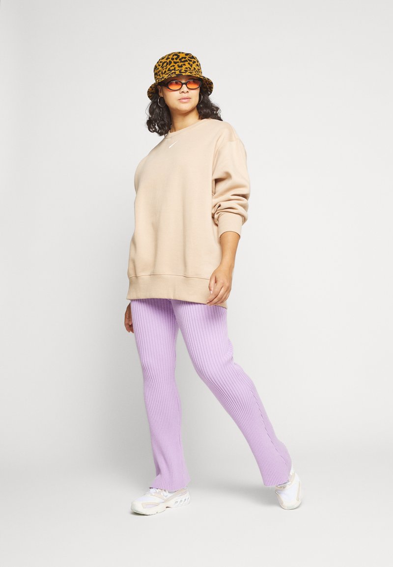 Sweatshirt beige surdimensionné, pantalon lavande côtelé, et chapeau seau à imprimé léopard. Des baskets blanches avec des détails rayés complètent le look.