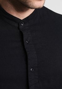 Camicia nera con colletto alla coreana, realizzata in un tessuto texturizzato, con una leggera lucentezza e quattro bottoni neri sul davanti.