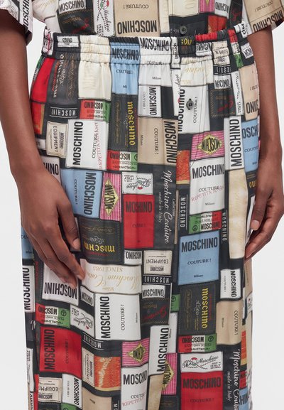 Les mains reposent sur une jupe avec un motif patchwork coloré composé de différentes étiquettes de la marque Moschino et blocs de texte.