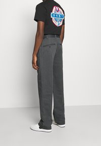 Zwarte t-shirt met kleurrijke "OBEY" grafische print op de achterkant, gecombineerd met grijze wijde broek. Schoenen zijn wit met zwarte accenten.