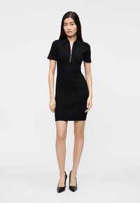 Črna obleka iz rebrastega pletenega materiala s kratkim ovratnikom z zadrgo in kratkimi rokavi, oprijetega kroja, združena z želodastimi črnimi petami.