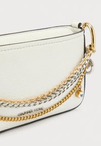 Λευκή τσάντα από δερμάτινη υφή με τρεις λουριά αλυσίδας σε ασημί και χρυσό, με ένα μικρό γούρι και μαύρες λεπτομέρειες στα άκρα.