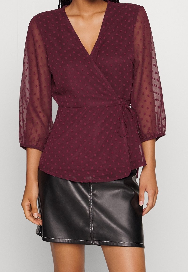 Blusa avvolgente borgogna con maniche lunghe trasparenti, texture a pois e vita con cravatta. Abbinata a una gonna in pelle nera con cuciture bianche.