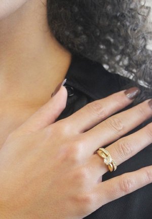 Anello d'oro con design intrecciato, caratterizzato da una pietra centrale trasparente e piccoli accenti trasparenti lungo la banda. Indossato su una mano con smalto scuro.