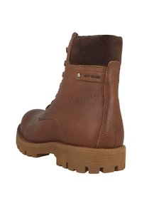 Brune læder ankelstøvle med en ruskindskrave, robust gummisål og metalakcent. Har en logoplade, tekstureret overflade og snøredetaljer.