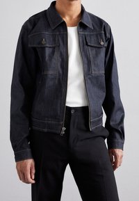 Hombre con chaqueta de mezclilla oscura y cremallera frontal sobre camiseta blanca de canalé y pantalones negros ajustados, contra un fondo liso.