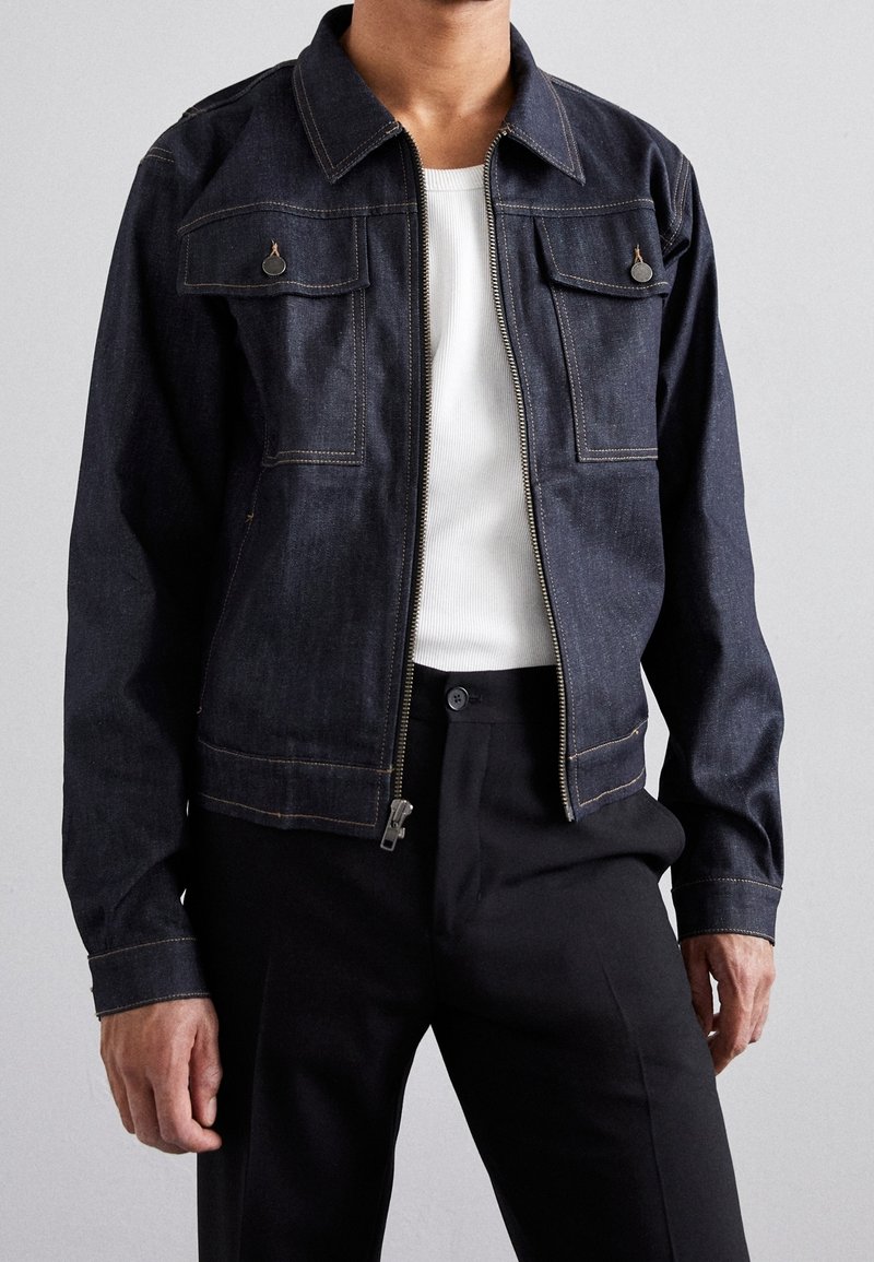 Hombre con chaqueta de mezclilla oscura y cremallera frontal sobre camiseta blanca de canalé y pantalones negros ajustados, contra un fondo liso.