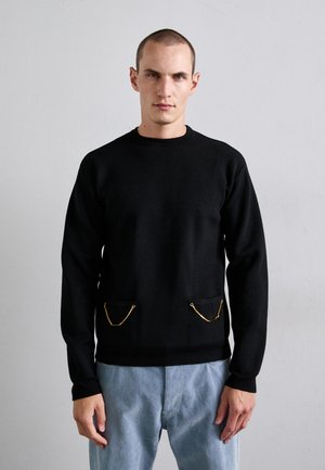 Junger Mann, der einen schwarzen Pullover mit goldenen Kettendetails an zwei Fronttaschen und hellblauen Jeans vor einem schlichten grauen Hintergrund trägt.