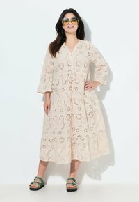 Helles Beige, langärmliges Kleid mit Lochstickerei und gestuftem Design. Abgeschlossen mit einer Knopfleiste. Kombiniert mit grünen Sandalen.