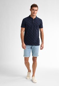 Camisa polo azul marino con patrón floral rojo, de mangas cortas y cuello. Combinada con pantalones cortos de mezclilla de un tono azul claro y zapatillas blancas.