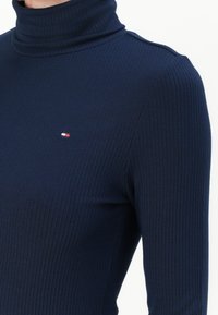 Marineblauwe geribbelde coltrui met lange mouwen en een klein logo-detail. De stof heeft een gladde textuur en een aansluitend ontwerp.