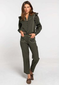 Combinaison à carreaux verts et noirs avec épaules volantées, boutons à l'avant et accents de poches contrastants. Stylisée avec des chaussures marron.