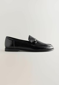Mocassin en cuir noir avec un petit talon et un détail boucle argentée sur le dessus, présenté de profil sur un fond uni.