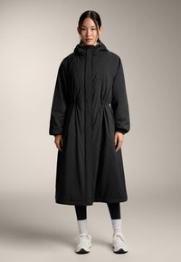 Fekete hosszú kapucnis kabátot, fekete leggingset, fehér zoknit és fehér sportcipőt viselő nő áll egy bézs háttér előtt.