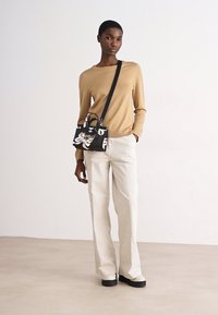 Borsa nera con design floreale bianco, tenuta da una persona che indossa una camicia a maniche lunghe color sabbia e pantaloni bianchi, in piedi su una superficie liscia.
