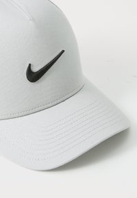 Jasnoszara czapka z wygiętym daszkiem, wykonana z gładkiego materiału. Posiada czarne haftowane logo Swoosh firmy Nike na przodzie.