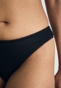 Črne teksturirane bikinijske hlačke z visokim izrezom, ki imajo širok, rebrast pas, kar poudarja njihov minimalističen stil.