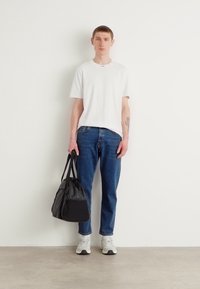 Bílý tričko s krátkým rukávem, modré džínové kalhoty a šedé sportovní boty. Drží černou tašku s dvěma popruhy. Minimalistický design, volný střih.