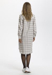 Robe à carreaux beige et marron, mi-longue avec des manches longues, dotée d'un dos boutonné et d'une coupe décontractée. Portée avec des collants noirs et des baskets blanches.