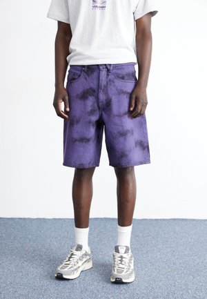Shorts de mezclilla morados con efecto tie-dye, cinco bolsillos, trabillas y corte recto. Combinados con zapatillas grises y calcetines blancos.