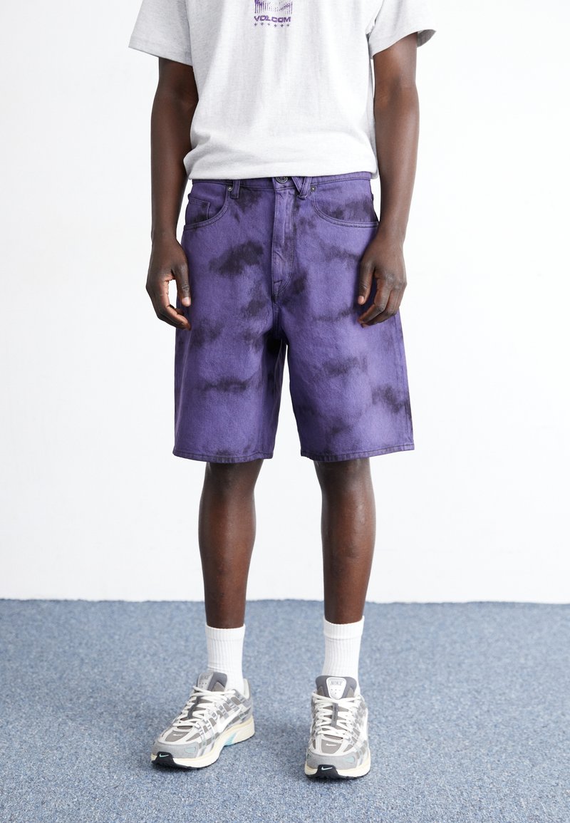 Shorts de mezclilla morados con efecto tie-dye, cinco bolsillos, trabillas y corte recto. Combinados con zapatillas grises y calcetines blancos.