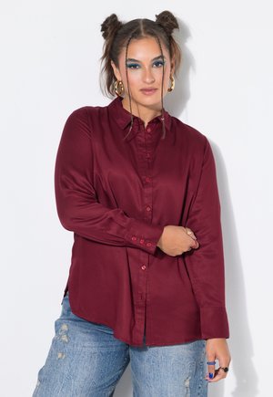 Kvinde med flettet hår og blå øjenskygge iført en burgundy knap-til-nederst skjorte og slidte blå jeans, poserende mod en hvid baggrund.