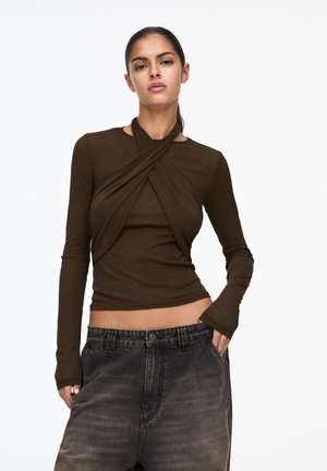 Bruine langemouwen top met een gedraaide voorkant. Heeft een aansluitende pasvorm en een zachte textuur. Gepaard met zwart vervaagde wijduitlopende jeans.