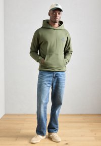 Olivovozelená mikina s kapucňou a predným vreckom, svetlomodré denimové nohavice a béžové tenisky. Model má na sebe smotanovú čiapku s zelenými akcentmi.