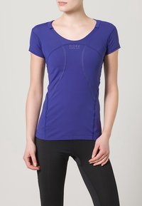 Maglietta tecnica viola a maniche corte con scollo a barca, cuciture accentuate e logo sul davanti, abbinata a leggings neri.