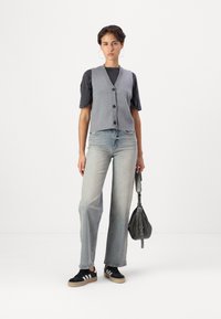 Gilet grigio a maglia con scollo a V e bottoni neri, indossato sopra una maglietta nera a maniche corte. Abbinato a jeans a gamba larga in denim chiaro e sneaker nere.