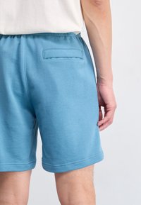 Blå sweatshorts tillverkade av mjukt tyg, med en bakficka, elastisk midja och en slät yta. Botten har dubbelsömmar.