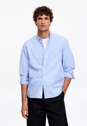 Mann mit lockigem Haar, der ein hellblaues Hemd über einem weißen T-Shirt und schwarze Hose trägt, steht mit einer Hand in der Tasche.