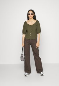Blusa verde-oliva de botões, com mangas bufantes, combinada com calças largas castanhas. Os acessórios incluem óculos de sol pretos e uma mala riscada.