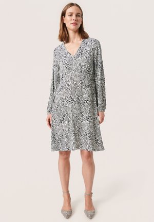 Blumenmusterkleid mit V-Ausschnitt, langen Ärmeln und ausgestellt geschnittenem Saum. Die Farben sind Weiß, Blau und Schwarz. Getragen mit grauen Absätzen.
