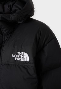 Melnā pūkā jacket ar izteiktu baltu logo kreisajā krūtīs, augsts apkakles un kapuce papildu siltumam.